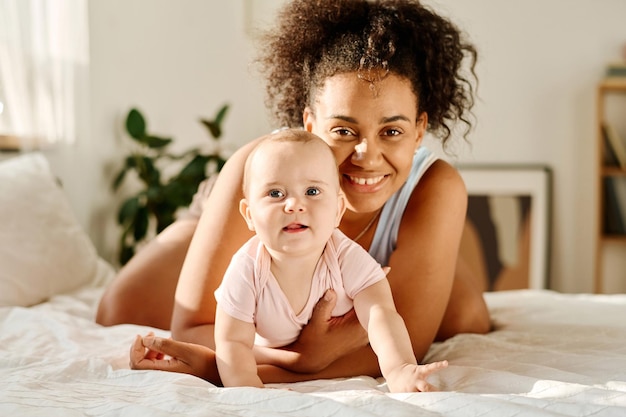 Ritratto di madre felice che sorride alla macchina fotografica mentre gioca con il suo bambino in camera da letto