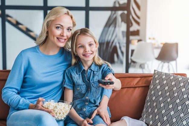 Ritratto di mamma felice con la figlia seduta sul divano che si abbraccia con popcorn che accende la tv