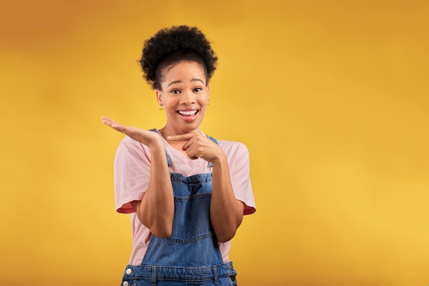 Ritratto di marketing e una donna di colore che punta il palmo della mano per la promozione di un prodotto su sfondo giallo in studio Sorriso pubblicitario o spazio con una felice giovane ambasciatrice del marchio femminile