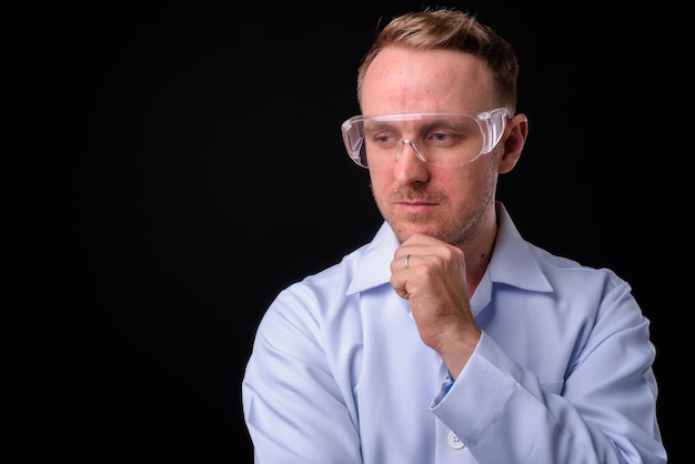 Ritratto di medico bello uomo biondo come scienziato con occhiali protettivi contro il muro nero
