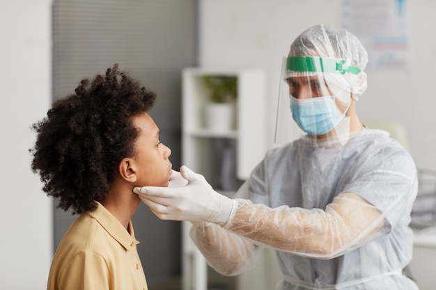 Ritratto di medico che indossa equipaggiamento protettivo completo che esamina ragazzo durante la consultazione in clinica, copia spazio