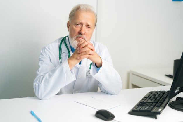 Ritratto di medico maschio adulto maturo serio in camice bianco con lo stetoscopio seduto alla scrivania con il computer in studio medico che guarda l'obbiettivo