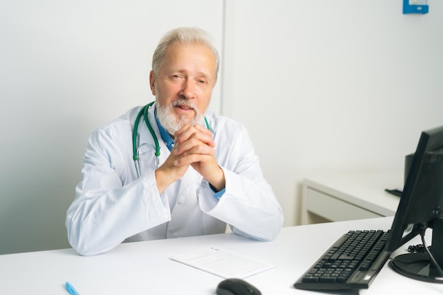 Ritratto di medico maschio adulto maturo sorridente in camice bianco con stetoscopio seduto alla scrivania con computer in studio medico guardando la fotocamera Medico senior fiducioso sul posto di lavoro in ospedale