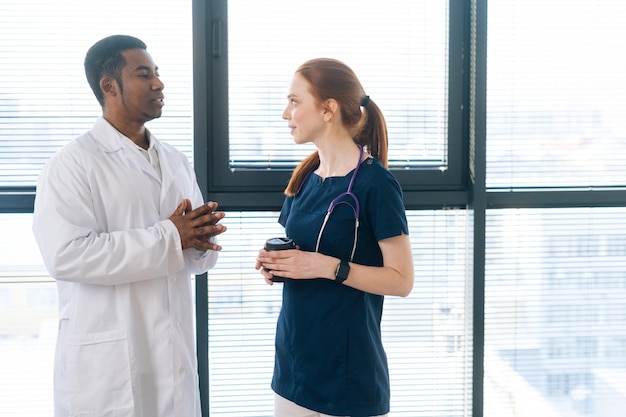 Ritratto di medico maschio nero afroamericano in camice bianco che parla con una giovane infermiera rossa positiva che indossa l'uniforme blu