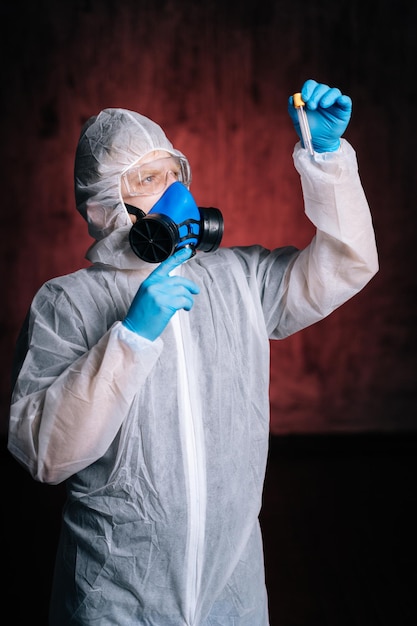 Ritratto di medico uomo in tuta protettiva, occhiali e respiratore tiene in mano la provetta e la guarda. Concetto di lavoro sul vaccino contro il coronavirus. Studio girato su sfondo rosso scuro isolato.