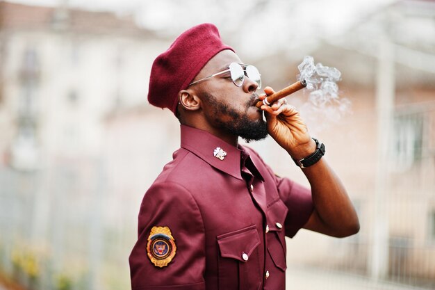 Ritratto di militare afroamericano in uniforme rossa, sungalasses e berretto. Sigaro di fumo del capitano.