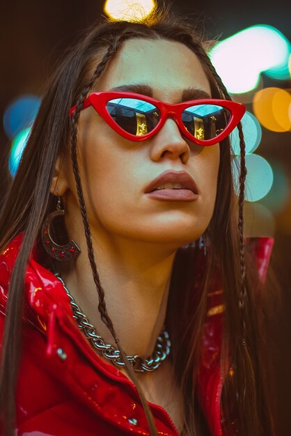 Ritratto di moda all'aperto di giovane donna glamour con i capelli intrecciati indossa piumino rosso e occhiali da sole rossi di moda sui lampioni al neon