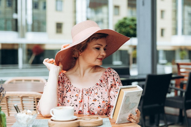 Ritratto di moda all'aperto di una splendida donna seduta in un bar Bevo caffè e leggo un vecchio libro una donna con un vestito e un cappello