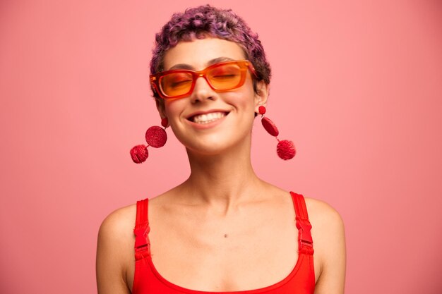 Ritratto di moda di una donna con un taglio di capelli corto con occhiali da sole colorati con accessori insoliti con orecchini sorridente su uno sfondo rosa brillante Foto di alta qualità