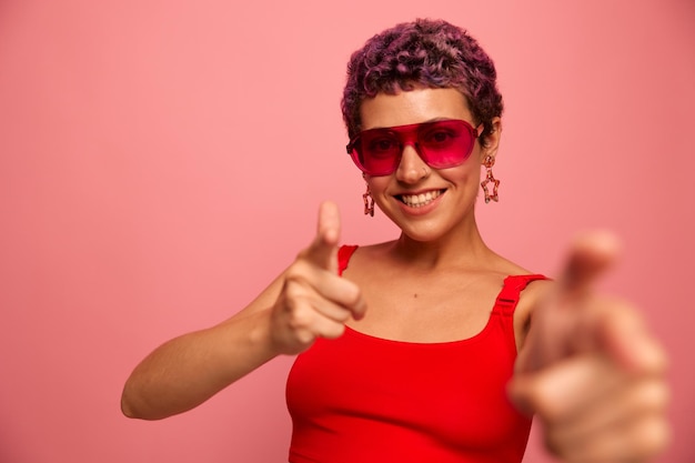 Ritratto di moda di una donna con un taglio di capelli corto in occhiali da sole colorati con accessori insoliti con orecchini sorrisi su uno sfondo rosa brillante mostra le mani alla fotocamera