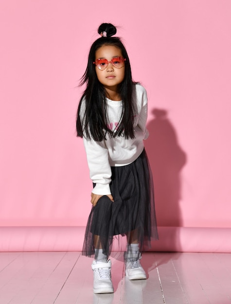 Ritratto di moda di una piccola modella coreana che posa professionalmente su uno sfondo rosa La ragazza indossa una gonna nera, maglione bianco, scarpe da ginnastica e occhiali rossi Concetto di moda per bambini