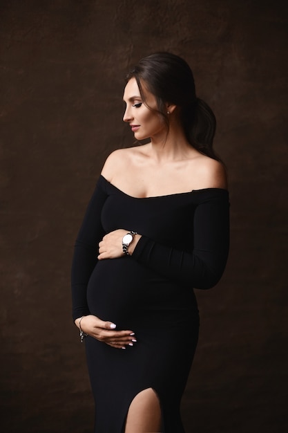 Ritratto di moda di una splendida modella incinta che indossa un abito da sera nero in posa su ba...