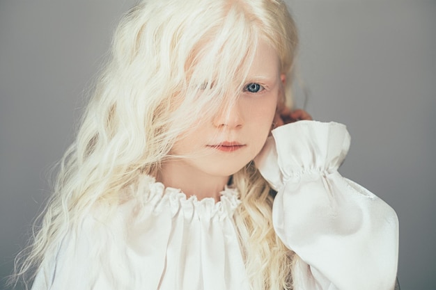 Ritratto di moda per bambini. Bellezza del bambino. Preziosa innocenza. Ritratto di dolce bambina bionda albina con capelli ricci occhi azzurri in camicetta bianca vintage isolata su sfondo grigio.