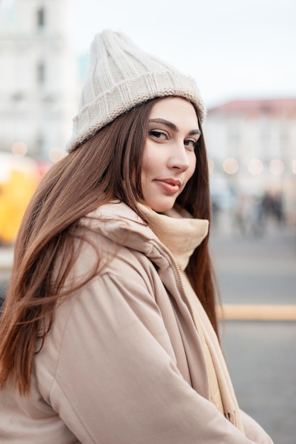 Ritratto di modello di donna abbastanza giovane in capispalla alla moda con cappello lavorato a maglia vintage e giacca beige in città