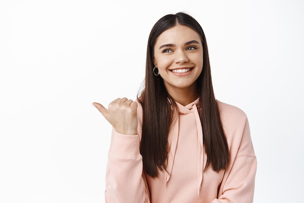 Ritratto di modello femminile bruna sorridente in felpa con cappuccio che punta e guarda a sinistra verso la pubblicità guardando felice il prodotto che mostra lo sfondo bianco del logo
