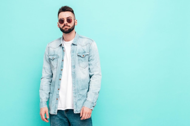 Ritratto di modello lambersexual bello e sicuro di sé hipster uomo vestito con giacca e jeans Moda maschile in posa vicino al muro blu in studio in occhiali da sole