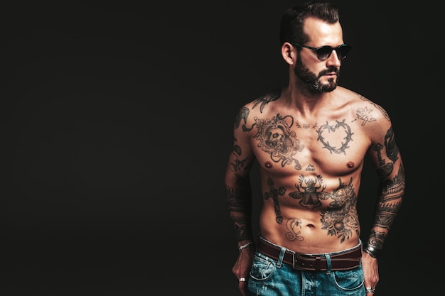 Ritratto di modello lambersexual bello fiducioso elegante hipster Uomo moderno sexy Torso nudo con tatuaggiUomo di moda in posa in studio su sfondo scuro in occhiali da sole