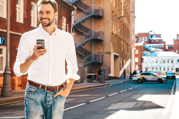 Ritratto di modello lambersexual bello fiducioso elegante hipster Uomo moderno vestito con camicia bianca Moda maschile in posa sullo sfondo della strada in occhiali da sole all'aperto al tramonto