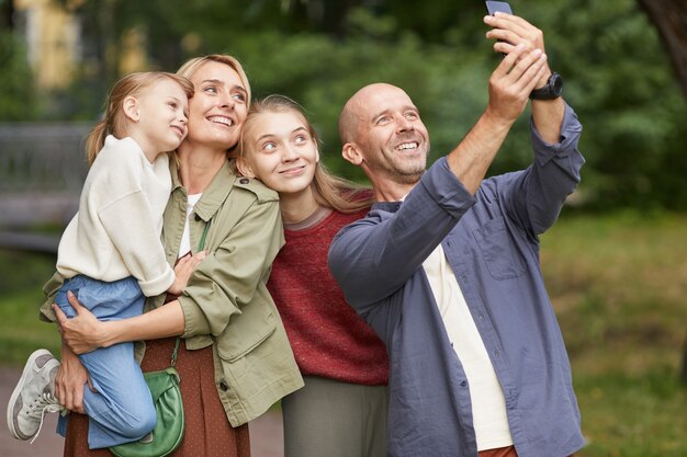 Ritratto di moderna famiglia felice con due figlie che prendono selfie foto all'aperto mentre vi godete una passeggiata nel verde del parco