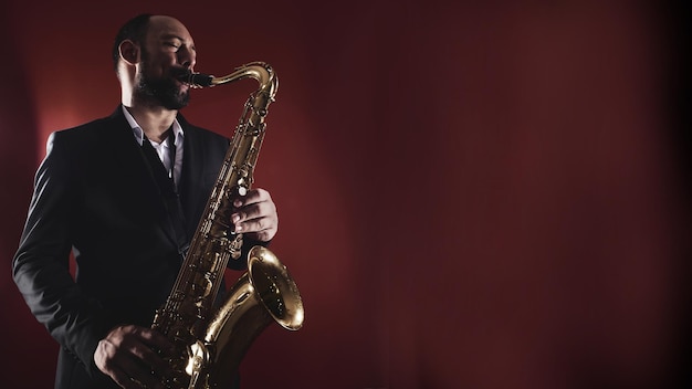 Ritratto di musicista professionista sassofonista uomo in tuta suona musica jazz sul sassofono, sfondo rosso in uno studio fotografico