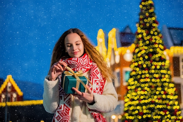 Ritratto di Natale di una bella ragazza in abiti caldi e una sciarpa in piedi sulla strada decorata per il nuovo anno sullo sfondo dell'albero di Natale con un regalo nelle sue mani.