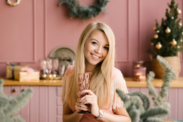 Ritratto di Natale di una ragazza con un vestito rosa e un cappotto di pelle di pecora sullo sfondo di un arredamento della cucina di Natale con un bicchiere di champagne Una donna si prepara a festeggiare il Natale e il nuovo anno
