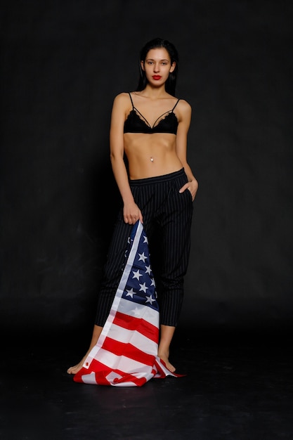 Ritratto di orgogliosa atleta femminile avvolto nella bandiera americana su sfondo nero. Giovane donna muscolare che guarda con sicurezza la macchina fotografica.