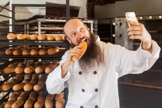 Ritratto di panettiere blogger adulto giovane affamato con barba lunga in uniforme bianca in piedi in fabbrica e fare selfie sugli scaffali con pane fresco sfondo pane gustoso concetto di professione al coperto