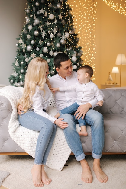 Ritratto di papà, mamma e figlio seduti per terra a casa vicino all'albero di Natale, tutti sorridono. Denim e camicie bianche. Natale in famiglia. Felicità.