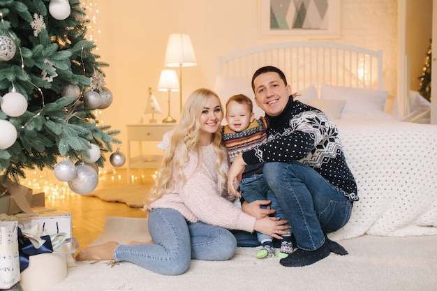 Ritratto di papà, mamma e figlio seduti per terra a casa vicino all'albero di Natale, tutti sorridono. Natale in famiglia. Felicità.