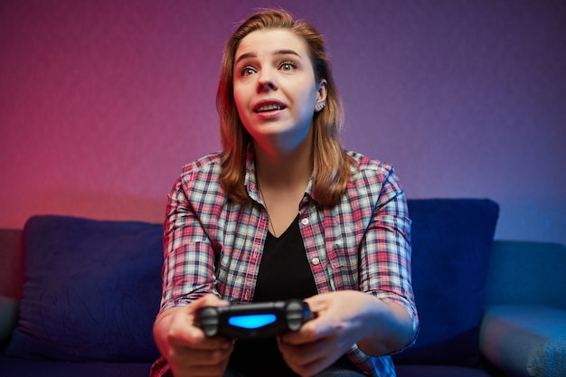 Ritratto di pazzo giocoso giocatore, ragazza che gode di giocare ai videogiochi al chiuso seduto sul divano, tenendo in mano il gamepad della console. Riposando a casa, buon fine settimana
