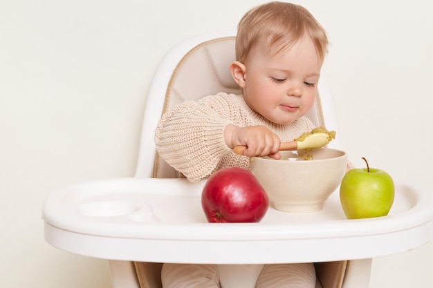 Ritratto di piccolo bambino carino e affascinante che mangia cibo da un piatto con un cucchiaio sullo sfondo mentre si siede con le mele intorno a mangiare il porridge per colazione