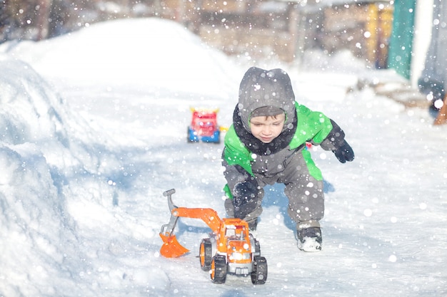Ritratto di piccolo bambino sveglio che si siede sulla neve e che gioca con il suo giocattolo giallo del trattore nel parco. Bambino che gioca all'aperto. Ragazzo felice con il giocattolo di costruzione. Concetto di stile di vita