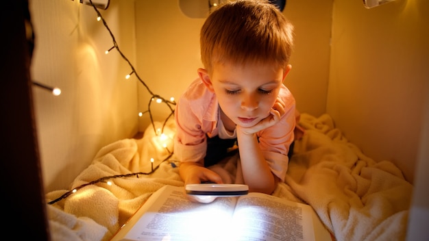 Ritratto di piccolo ragazzo intelligente che legge un grande libro di storia di notte. Bambino che gioca nella casa di cartone giocattolo. Concetto di educazione dei bambini e lettura in camera oscura.