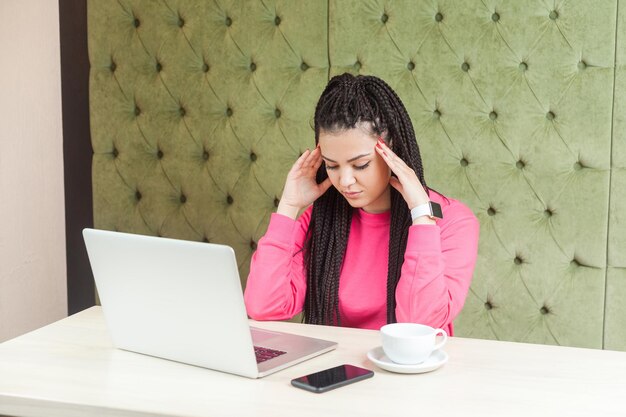 Ritratto di premurosa e triste giovane ragazza freelance con l'acconciatura nera dei dreadlocks seduta e lavorando sul laptop tenendo la testa verso il basso e cercare di ricordare le informazioni riprese in studio al coperto