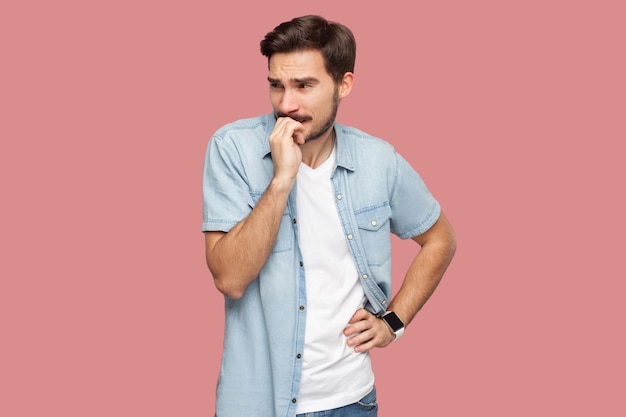 Ritratto di preoccupazione nervosa bel giovane barbuto in camicia blu stile casual in piedi guardando lontano e mangiandosi le unghie. girato in studio al coperto, isolato su sfondo rosa.