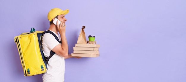 Ritratto di profilo del fattorino in berretto e maglietta bianca che parla al cellulare con il cliente che tiene la pizza in una scatola di cartone e caffè isolato su sfondo viola