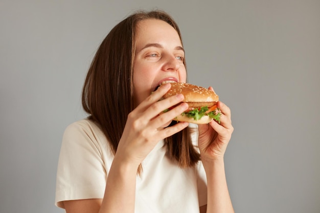 Ritratto di profilo di una donna caucasica dai capelli castani che morde un delizioso hamburger appetitoso essendo estremamente affamata godendosi cibo spazzatura dieta finale con pasto cheat