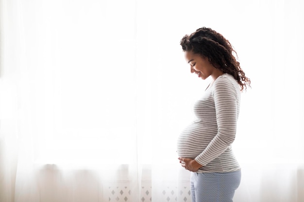 Ritratto di profilo di una donna incinta sorridente nera che abbraccia teneramente la pancia a casa