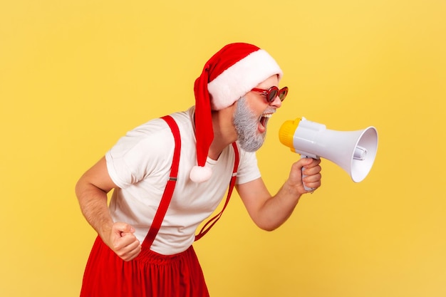 Ritratto di profilo eccitato uomo barbuto grigio nervoso in cappello di Babbo Natale che urla tenendo il megafono dell'altoparlante, divertendosi a celebrare le vacanze invernali. Studio indoor girato isolato su sfondo giallo