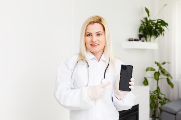 Ritratto di profilo laterale sorridente dottoressa, professionista sanitario in camice bianco con stetoscopio, analizzando i risultati dei dati sullo smartphone mobile in piedi nel corridoio del corridoio dell'ospedale