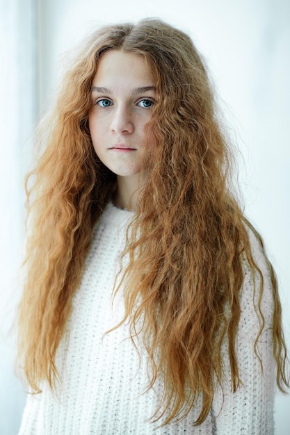 Ritratto di ragazza adolescente con gli occhi azzurri con lussuosi capelli rossi ondulati lunghi in piedi vicino alla finestra nella luce del giorno invernale. Lentiggini adorabili. Mondo interiore. Capelli morbidi e arruffati. Bellezza naturale. Luce naturale.