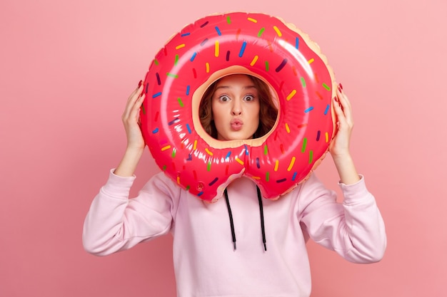 Ritratto di ragazza adolescente dai capelli ricci giocosa in felpa con cappuccio che tiene anello di gomma a ciambella dietro il viso e labbra imbronciate, divertendosi. Studio indoor girato isolato su sfondo rosa