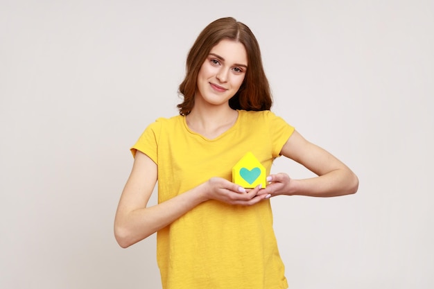 Ritratto di ragazza adolescente in maglietta gialla che tiene in mano vicino al cuore piccola casa di carta che guarda l'obbiettivo con un sorriso piacevole, sognando la propria casa. Studio indoor girato isolato su sfondo grigio.