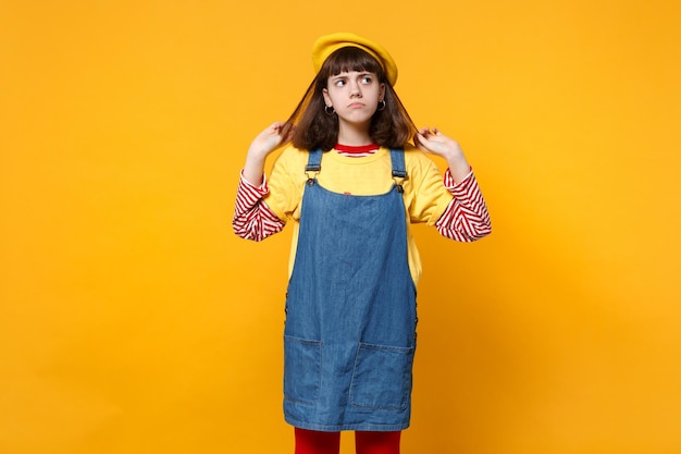 Ritratto di ragazza adolescente pensierosa in berretto francese, prendisole in denim che tiene i capelli, guardando in alto isolato su sfondo giallo muro in studio. Persone sincere emozioni, concetto di stile di vita. Mock up spazio di copia.