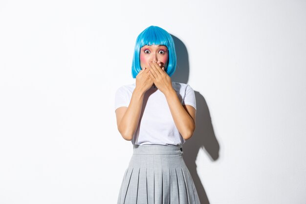Ritratto di ragazza asiatica scioccata in parrucca blu senza fiato per lo stupore, coprire la bocca con entrambe le mani e fissare la telecamera, in piedi.