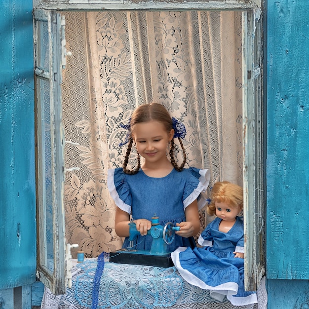 Ritratto di ragazza bambino sarto cucire fare vestiti per bambole su una macchina da cucire per bambini nella finestra di