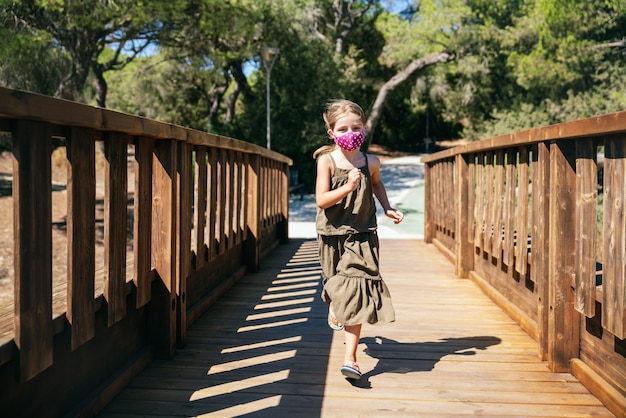 Ritratto di ragazza bionda con gli occhi azzurri con maschera facciale in vacanza che attraversa un ponte di legno con alberi di pino con abito verde nel mezzo della pandemia di coronavirus