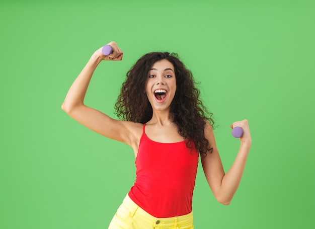 Ritratto di ragazza bruna che indossa abiti estivi facendo sport e sollevando pesi contro il muro verde