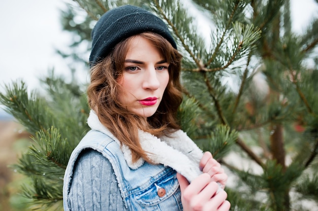 Ritratto di ragazza bruna in giacca di jeans e cappello all'albero di Natale.